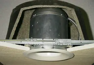 A recessed ceiling light fixture with housing partially installed in a cutout section of drywall, compatible with high hat covers for added protection and energy efficiency.