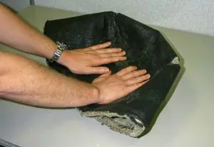 Two hands pressing down on a thick, black, flexible in-ceiling speaker cover placed on a light-colored table.