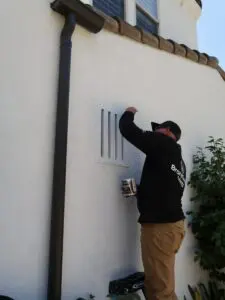 Man in black jacket installs vent cover on white houses exterior wall.
