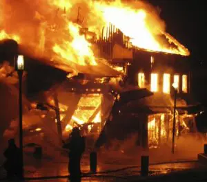 A large building engulfed in bright flames at night, with firefighters silhouetted in the foreground.