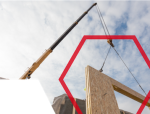 A crane lifts a wooden wall panel at a construction site under a partly cloudy sky.