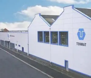 White industrial building with blue trim, large windows, and TENMAT logo on the side under a partly cloudy sky.