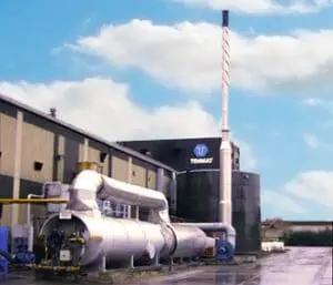 Industrial building with large metal pipes and a tall smokestack under a partly cloudy sky.