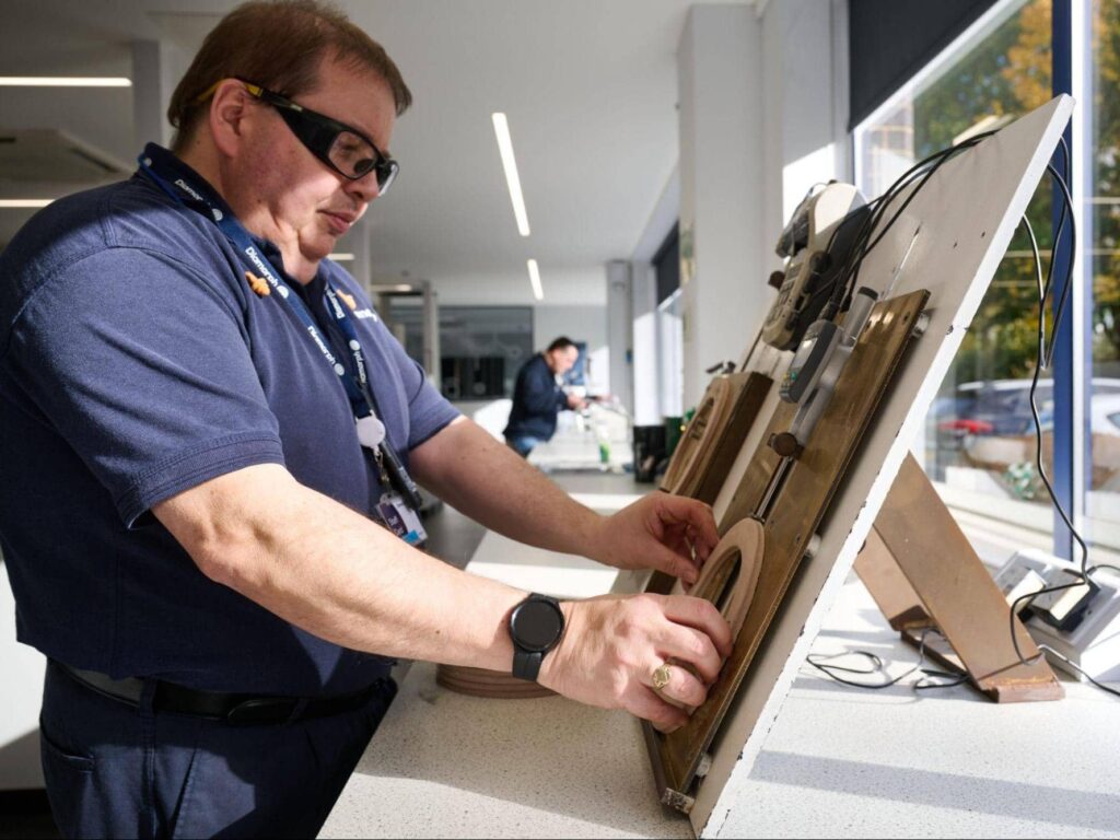Man with spectacles works on a round wooden tool at a counter in a bright room.